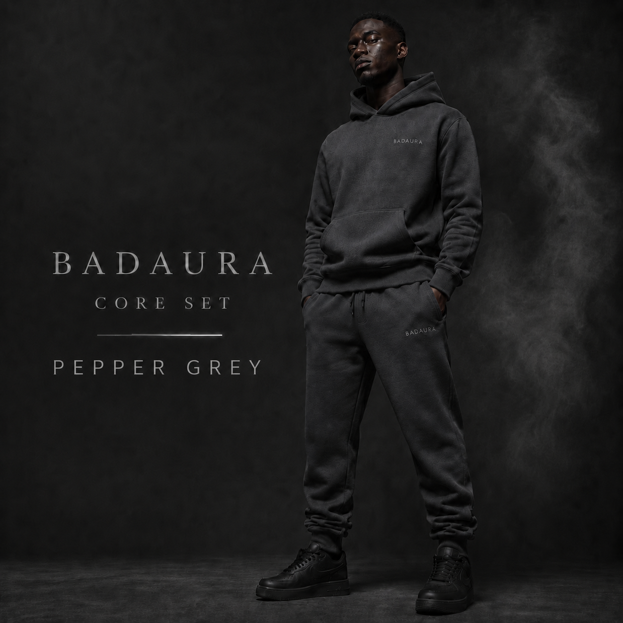 Person wearing a dark gray hoodie and pants with 'Badaura' branding on a black background.